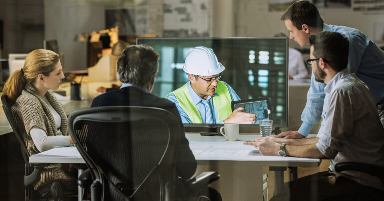 A group of safety professionals gather around a table while a large monitor shows a man in a hard hat demonstrating a procedure