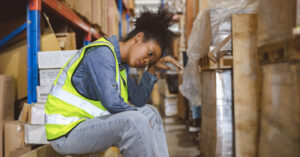 A woman in a neon vest sits down on a box between rows of warehouse shelves, touching her forehead with the back of her hand as she experiences high heat.